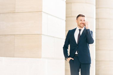 Attractive male CEO solves working issues with business partner during phone conversation, dressed in formal black suit, keeps hand in pocket, looks happily into diastance. Business and people