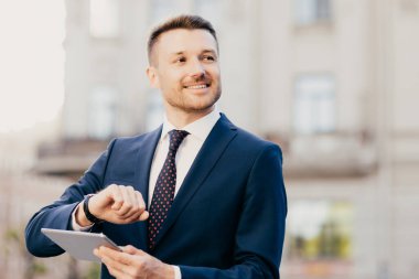 Attractive male business owner in elegant black suit, looks at wristwatch, holds modern tablet, messages with friends and colleagues in social networks, comes on meeting too early. Career concept