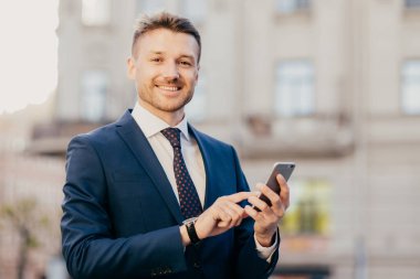 Satisfied businessman holds smart phone, reads email from investor, happy to recieve good news about business company, stands in urban territory, wears black suit and elegant tie. Technology concept