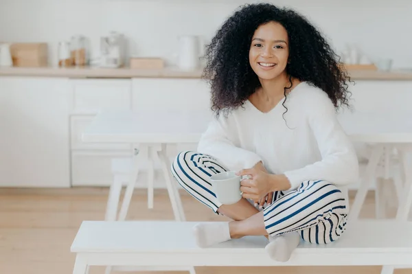 Smiling relaxed African American female sits crossed legs on bench against kitchen interior, wears white sweater and striped pants, drinks hot beverage, enjoys domestic atmosphere. Coffee time