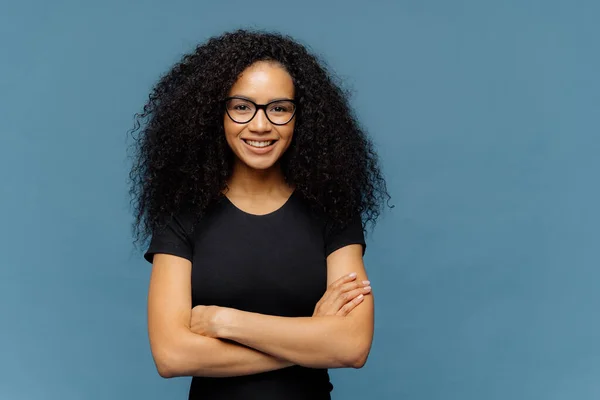 腰朝上拍摄的微笑的非洲裔美国妇女有双臂折叠 戴着眼镜和休闲的黑色T恤 享受与对话者愉快的交谈 在蓝色工作室墙上摆姿势与空白空间 — 图库照片