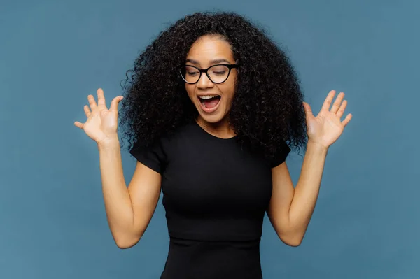苗条的黑皮肤女人举手 张开嘴 手势积极从积极的情绪 集中下来 穿着休闲的黑色T恤 孤立在蓝色背景 种族概念 — 图库照片