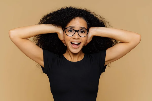 Young woman with curly Afro hair covers ears, shouts as demands be quiet, cant stand noise, keeps mouth opened, wears spectacles and casual black t shirt, isolated over brown studio background.