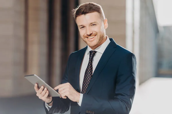 Photo of successful male banker does remote work on digital tablet, has happy expression, dressed in formal black suit, has happy look at camera. Positive young man makes transactions online
