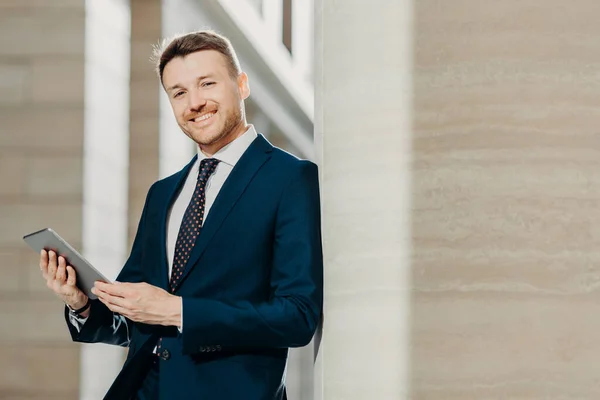 Portrait of stylish male entrepreneur has cheerful expression, holds digital tablet, reads financial news on web page, uses free internet connection, wears formal suit and tie. Career concept