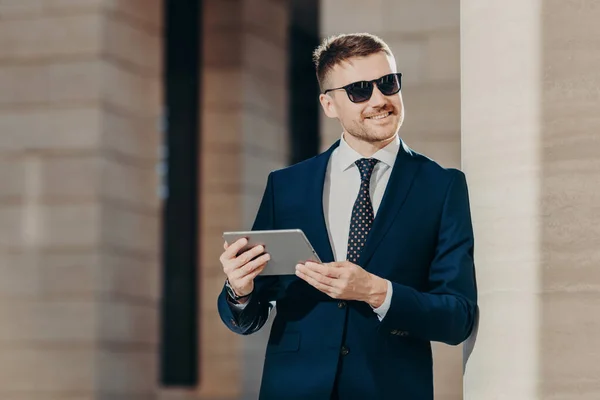 Positive smiling male manager with stubble, wears sunglasses and black suit, holds modern touch pad, checks email in internet, recieves positive news, looks into distance. People, business and style