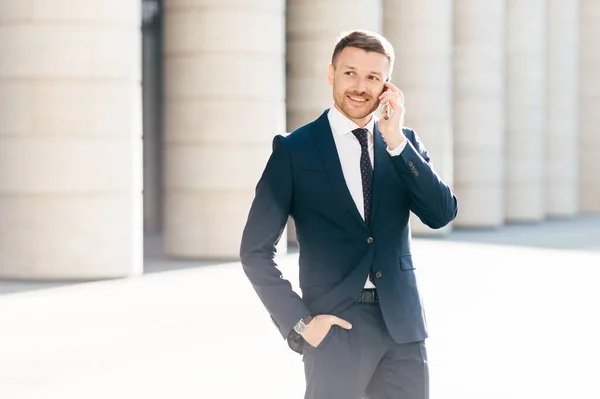 Portrait of successful male manager satisfied with mobile tariffs, makes phone call, uses banking service operator, wears formal suit, stands in urban setting outsie. Professional man worker