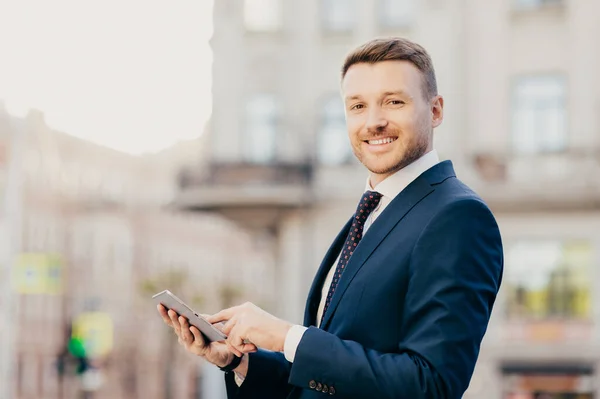 Sideways portrait of successful male freelancer works distantly on touchpad, stands outdoor, being in good mood after having dinner in luxury restaurant with colleague, connected to wireless internet