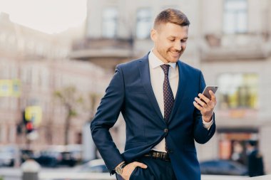 Young succesfful delighted happy businessman reads text message on modern cellular, wears elegant suit, holds hand in pocket, enjoys online communication. People, business, technology concept