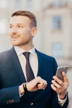 Attractive male comes on meeting with client or partner who is late, looks at watch and holds smart phone, looks aside, hopes to see someone into distance, dressed in formal suit, stands outdoor