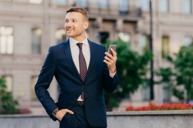 Happy attractive male financier in elegant wear, uses modern smartphone for following financial news on special installed application, stands on street, looks aside with cheerful expression.