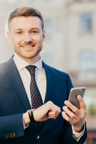 Smiling manager director with watch on arm, dressed elegantly, holds smart phone, reads positive news about his new project, poses outdoor, waits for colleague near office building. Job concept