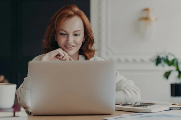 Experienced red haired businesswoman reads publication on modern laptop computer, concentrated at screen with pleasant smile, makes research for planning startup, checks mail box, works from home