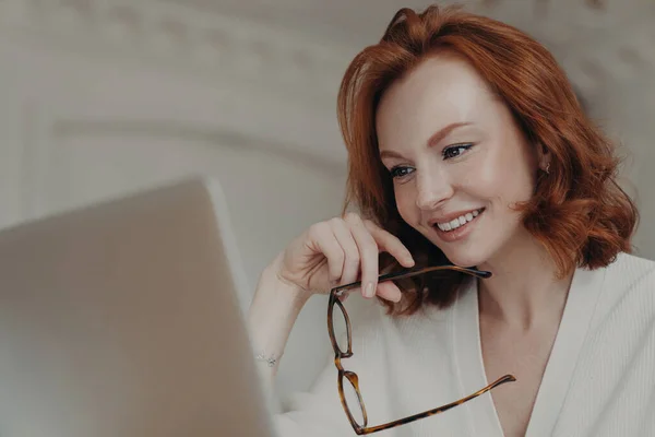 プロの笑みを浮かべて生姜の女性のウェブデザイナーは プロジェクトの仕事を作成するためのラップトップコンピュータ上のアプリケーションを使用して オンライン研究やインターネットを閲覧し 眼鏡を保持し 幸せな表情をしています — ストック写真