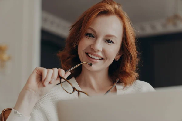 Cheerful redhead female web designer works on freelance, poses at opened laptop computer, holds glasses, shares content in social networks, smiles gently, poses at home. People and technology