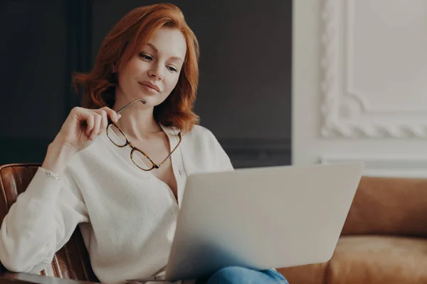 集中深刻な赤毛の女性は屋内でリモートジョブを行い 快適なアームチェアに座って 透明なガラスを保持し インターネットのウェブサイト上で出版物を読み取り 家庭用無線Lanを使用しています 労働時間 — ストック写真
