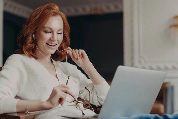 Satisfied happy foxy successful woman has video conference, uses modern laptop computer and earphones, browses data and works on remote project, poses in modern apartment, watches online webinar