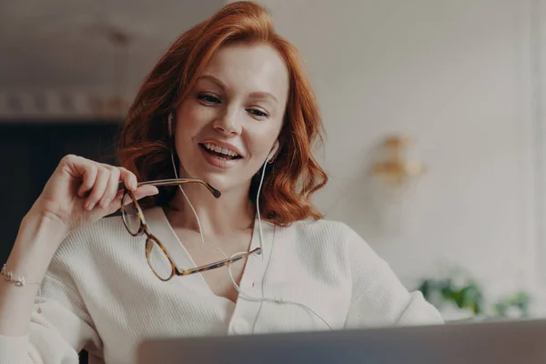 自信を持って満足した赤毛の女性は ラップトップコンピュータに集中し ラップトップコンピュータ上のビデオや映画を見て フレンドリーなビデオ通話を楽しんで 透明なガラスを保持し 無料のインターネット接続を使用しています — ストック写真