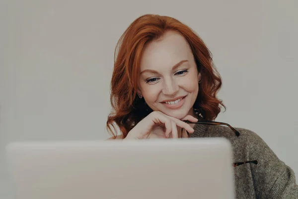 Successful female student with ginger hair and pleasant smile watches interesting tutorial video or webinar, uses laptop computer for e learning, reads information online, keeps hand under chin