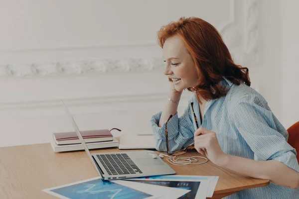 Profile shot of pleased redhead woman entrepreneur enjoys video conference with colleague, poses at desktop with paper documents, pleased to work from home, glad to see friend. Vitrual reality