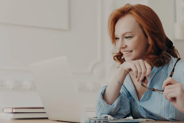 赤頭の水平ショットヨーロッパの女性は ノートパソコンの画面に焦点を当て 自宅のオフィスでポーズ 眼鏡を保持し コンピュータ上のビデオを見て 良いニュースを楽しんで シャツを着て 距離学習を楽しんで — ストック写真
