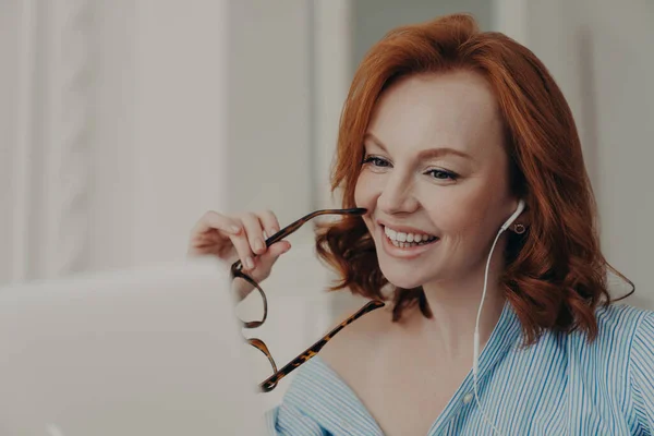Beautiful cheerful redhead woman uses laptop computer for online conference, talks with colleagues distantly, holds spectacles, discusses working issues, works freelance from home. Technology concept