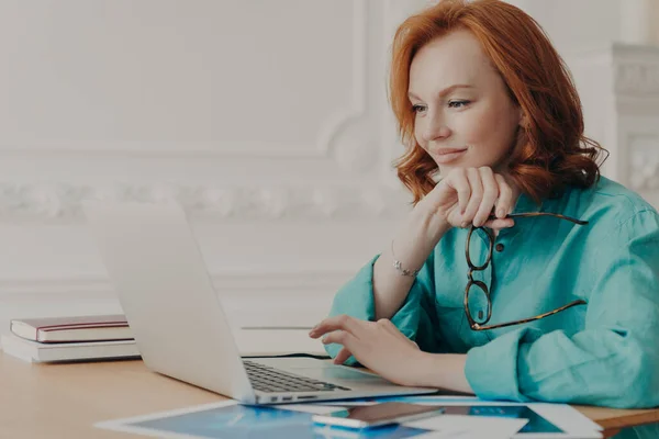 Busy Redhead Woman Entrepreneur Works Modern Laptop Holds Spectacles Poses — Stock Photo, Image