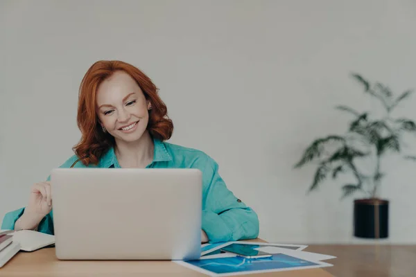 幸せな生姜の女性のフリーランサーの水平ショットは 自宅でラップトップコンピュータ上で動作します 研究オンライン ソフトウェアを使用します インターネット上のサーフ情報 デスクトップに座っています 積極的に笑顔 リモートで動作します — ストック写真