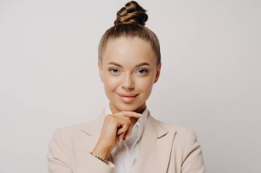 Kendine güveni tam, bej takım elbiseli genç bir iş kadını. Gri arka planda elini çenesinin altında tutarken kendine güveni tam. Kar veya iş hakkında düşünürken ileriye bakıyor.