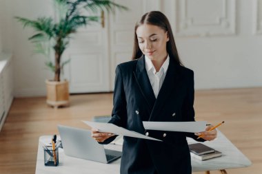 Zarif klasik kıyafetli kendine güvenen genç iş kadını belgelere bakıyor ve iyi proje sonuçlarıyla mutlu oluyor, gülümseyen kadın yönetmen modern ofiste duruyor ve çalışma gününün tadını çıkarıyor.