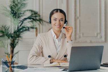 Resmi takım elbiseli, mikrofonlu bir kulaklık takmış müşteriye gülümseyen genç bayan müşteri destekçisi portresi. Modern ofiste iş kadınının iş gününün keyfini çıkarması.