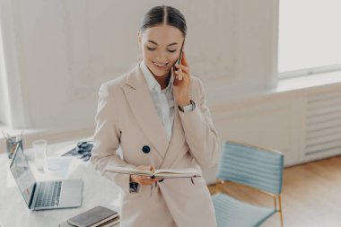 Çekici genç iş kadını patronla iş meseleleri hakkında konuşurken iş yerinin yanında dizüstü bilgisayarla şık bir ofiste durup not defterine bakıyor.