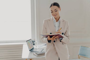 Kendine güvenen genç iş kadını modern ofisin şık iç kısımlarında duruyor, gülümseyen profesyonel bayan girişimci bej takım elbise giyiyor çalışma saatlerini kalem ile not ediyor.