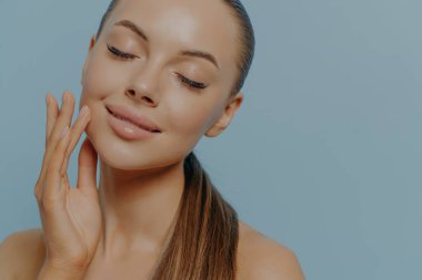 Pure natural beauty. Closeup portrait of young pleased woman with closed eyes touching soft healthy skin after skincare treatment or spa procedure, posing with bare shoulders on blue background