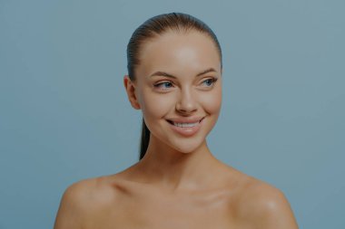 Skincare and beauty. Young attractive woman with natural makeup and healthy glowing skin looking aside and smiling while posing shirtless on blue background. Facial care and wellness