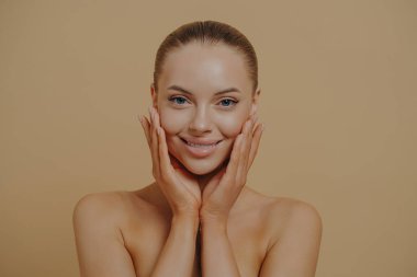 Cosmetology and dermatology. Headshot of beautiful woman touching cheeks, enjoying her soft and smooth skin, standing half-naked over beige background. Face of smiling european lady without make-up