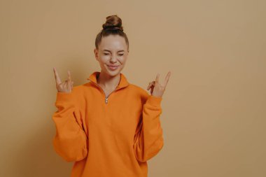 Yeah, i did it. Overjoyed young woman isolated on beige studio background showing rock-n-roll gesture, smiling with closed eyes, being proud of success, excited female making sign of horns