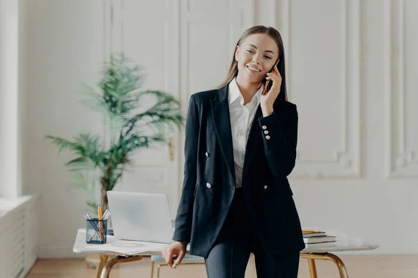 オフィスで働く黒の正式なスーツの若いプロの陽気なビジネスの女性は クライアントと電話で話し 開いたラップトップで仕事のテーブルにもたれながら 喜んで笑顔 オフィスに立って — ストック写真