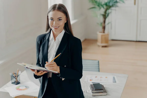 Portrait of young successful business consultant in black formal suit holding document, looking forward and smiling at camera in front of white desk at big light office. Business people at work