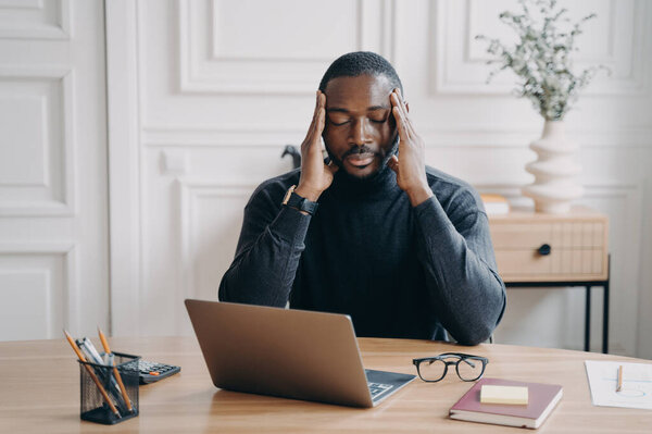 Concerned young african american man employee suffering from headache at workplace, sitting with closed eyes and frustrated face expression, worried male office worker thinking of problem solution