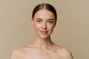 Indoor shot of beautiful young woman with combed hair stands half naked indoor has natural beauty isolated over beige background. Refreshed female model has healthy skin after spa treatments