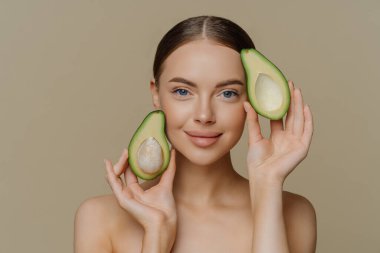 Tişörtsüz kadın modelinin fotoğrafı kameraya nazik bir ifadeyle bakıyor. Yüzüne yakın avokado yarımları bej arkaplanda izole edilmiş kozmetik tedavilerden sonra sağlıklı bir cilde sahip..