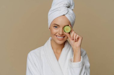 Mutlu ve yenilenmiş bir kadının yatay pozu, cildine bir parça salatalık katar. Duştan sonra banyo elbisesi giyip gülümseyerek organik ürünler kullanır..