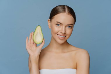 Doğal cilt bakımı kavramı. Güzel esmer genç kadın avokadonun yarısını elinde tutuyor organik kozmetik ürünlerden hoşlanıyor güzellik bakımı için gerekli yağlardan hoşlanıyor mavi arka planda banyo havlusuna sarılı pozlardan hoşlanıyor