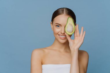 Kadınların cilt bakımı doğal kozmetik ürünleri ve sağlıklı yaşam tarzı konsepti. Memnun bir genç kadın, gözlerini avokadoyla kaplıyor. Cilt bakımını seviyor. Banyo havlusuna sarılmış.