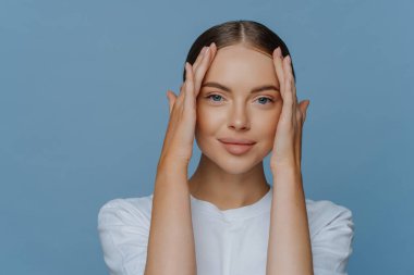Çekici, genç, esmer kadın modelin yüzüne dokunması cildin yumuşaklığından zevk alır. Günlük beyaz tişört giymiş doğal bir makyajı vardır. Mavi arka planda sakince izole edilmiş kameraya bakar. Güzellik konsepti
