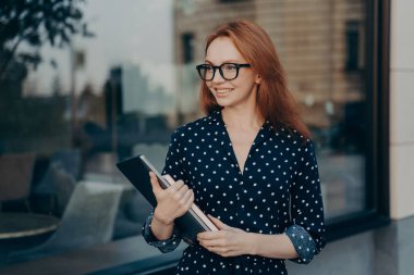 Kızıl saçlı başarılı bir iş kadını modern bir cihaz taşır ve not defteri resmi toplantıya gider. Gözlük takar, siyah benekli elbise giyer. Açık havada restoranın yakınında mutlu bir şekilde poz verir.