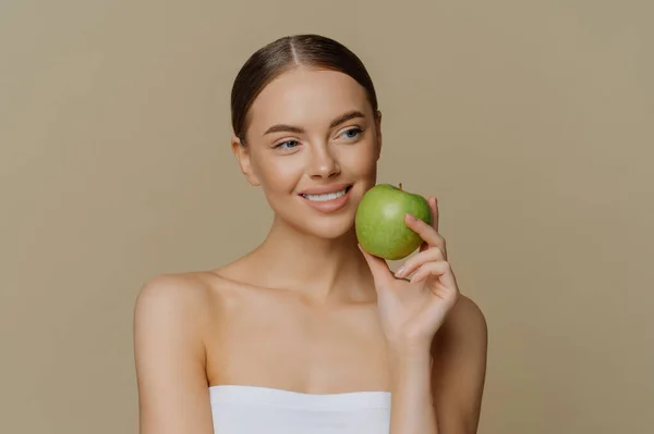 魅力的なヨーロッパの女性は顔の近くにリンゴを保持しています笑顔優しく白い完璧な歯を持っています健康的なきれいな肌シャワータオルスタンドに包まれ茶色の背景に裸の肩で — ストック写真