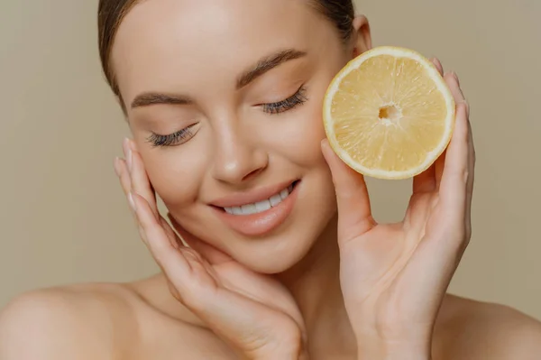Headshot of sensual charming young woman wears minimal makeup touches face gently holds slice of lemon cares about freshness of skin has citrus peeling stands half naked against brown background