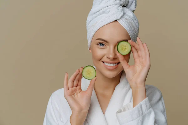 Skin care natural beauty and cosmetology concept. Positive refreshed beautiful woman holds slices of fresh cucumber makes mask for face skin dressed in domestic robe wrapped bath towel on head